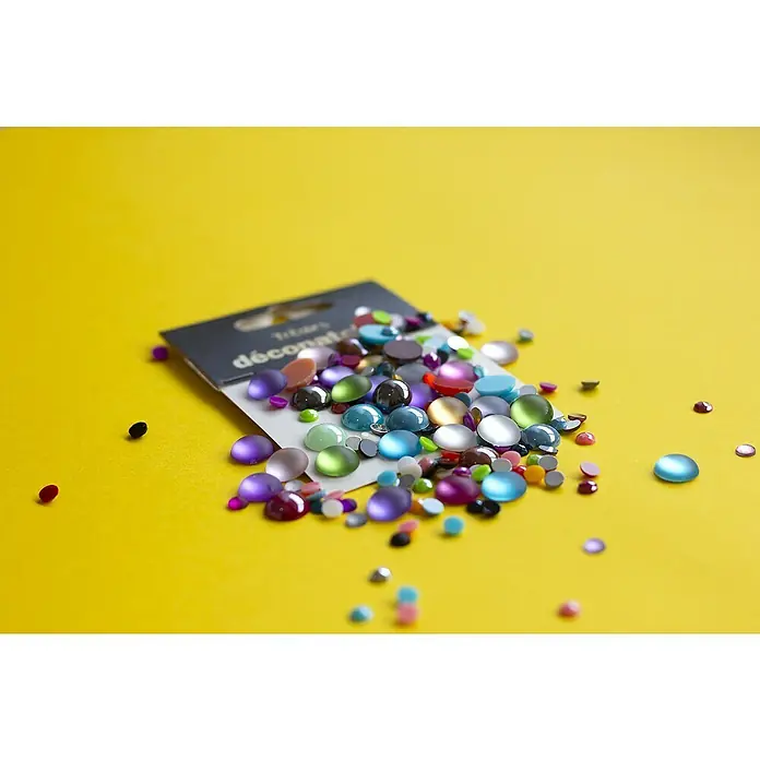 Décopatch Pieza decorativa Trésors Flashy - 24 ud., 1 x 1 cm - Primer plano de piedras decorativas de colores esparcidas sobre un fondo amarillo, perfectas para proyectos de manualidades y decoración.