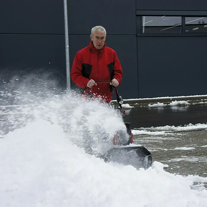 AL-KO 36V Akku-Schneefräse ST4048 - 36 V, Ohne Akku, Arbeitsbreite: 48 cm - Ein Mann benutzt eine Schneefräse, um Schnee von einer gepflasterten Fläche vor einem Gebäude zu räumen. Die Schneefräse wirbelt den Schnee hoch.