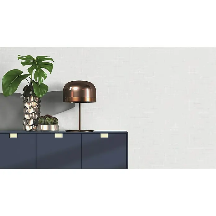 Rasch Wallton Vliestapete Premium Struktur I - Weiß, Uni, 10,05 x 0,53 m - Elegante Kommode in Dunkelblau mit goldenen Akzenten, darauf eine Lampe, eine Monstera-Pflanze und ein Kaktus. Stilvolle Möbel für ein modernes Zuhause.