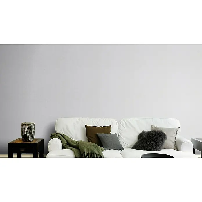 Rasch Bambino XIX Vliestapete - Blaugrau, Uni, 10,05 x 0,53 m - Ein weißes Sofa mit grüner Decke und dekorativen Kissen, darunter ein Fellkissen, vor einer grauen Wand. Beistelltisch mit Vase links.