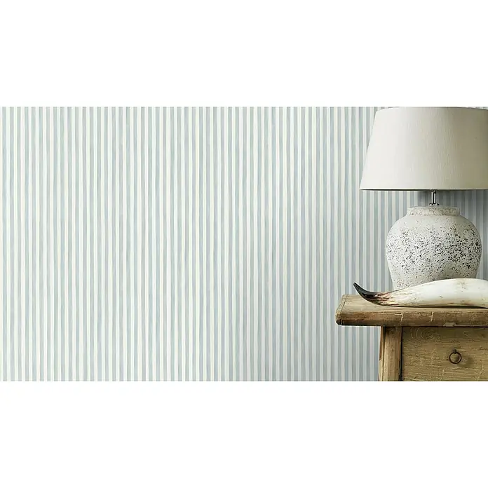 Rasch Bambino XIX Vliestapete - Blau, Streifen, 10,05 x 0,53 m - Nahaufnahme einer Tischleuchte mit cremefarbenem Schirm auf einem Holztisch vor einer gestreiften Tapete in Blau und Weiß.
