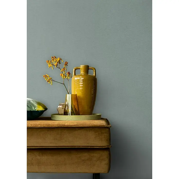 Rasch Salsa Vliestapete TextilstrukturPetrolgrau, Uni, 10,05 x 0,53 m - Gelbe Vase mit Griffen und Blumen auf einem braunen Samthocker vor einer blaugrauen Wand.