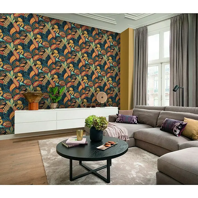 Rasch Salsa Vliestapete - Ocker, Uni, 10,05 x 0,53 m - Elegantes Wohnzimmer mit einem grauen Ecksofa, einem schwarzen Couchtisch und einer auffälligen Blättertapete.
