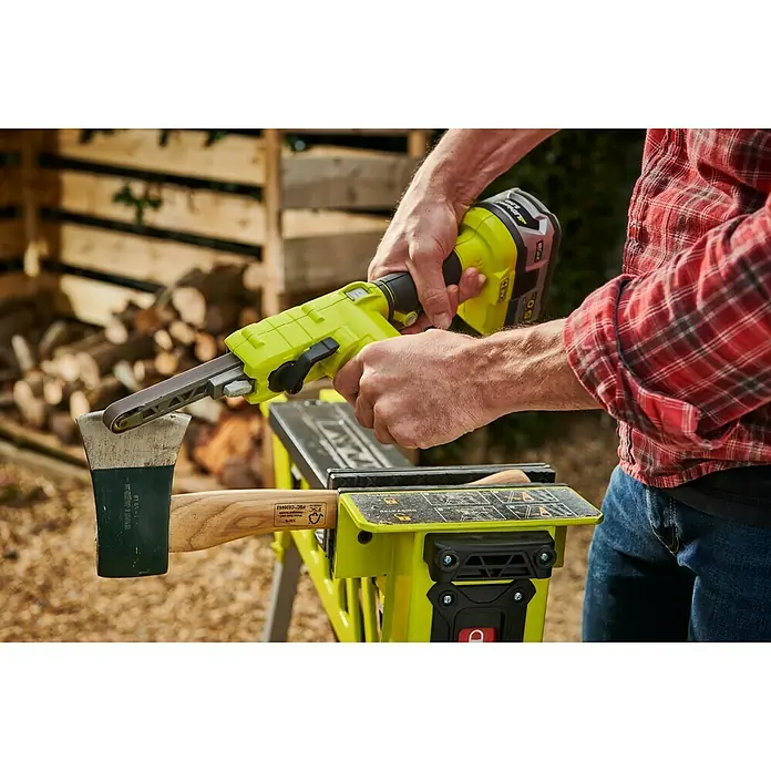 Ryobi ONE+ Maschinenpaket - Akku-Bandschleifer R18PF-0 + gratis Starter-Kit - Eine Person schärft eine Axt mit einem Ryobi-Bandschleifer auf einer Werkbank.