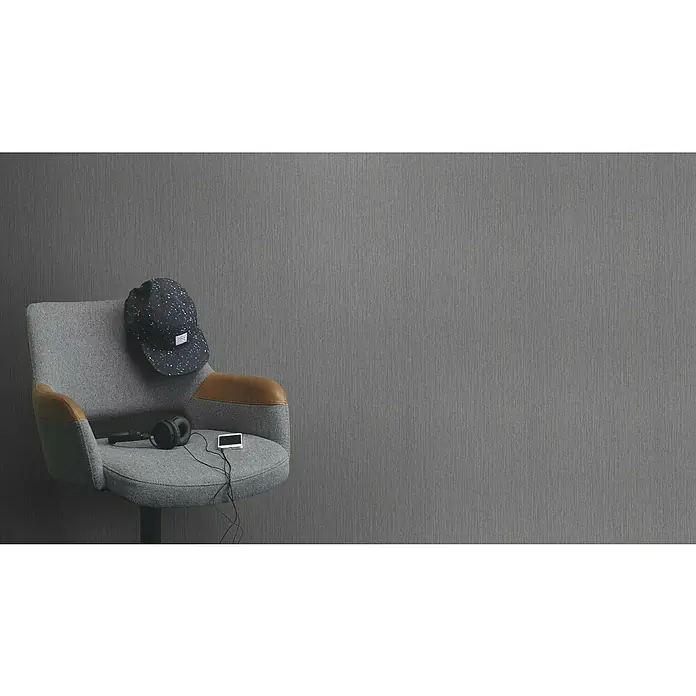 Rasch Florentine III Vliestapete Textile Stoffoptik - Dunkelgrau, Uni, 10,05 x 0,53 m - Ein grauer Sessel mit Lederakzenten, Kopfhörern, einer Mütze und einem Musikplayer vor einer grauen Wand.