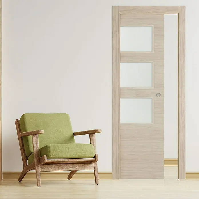 Puerta corredera de madera acristalada Arizona - 72,5 x 203 cm, Roble arena, Maciza, Con uñero - Puerta de madera clara con tres inserciones de vidrio cuadradas junto a un sillón verde. La puerta está parcialmente abierta, revelando el interior.
