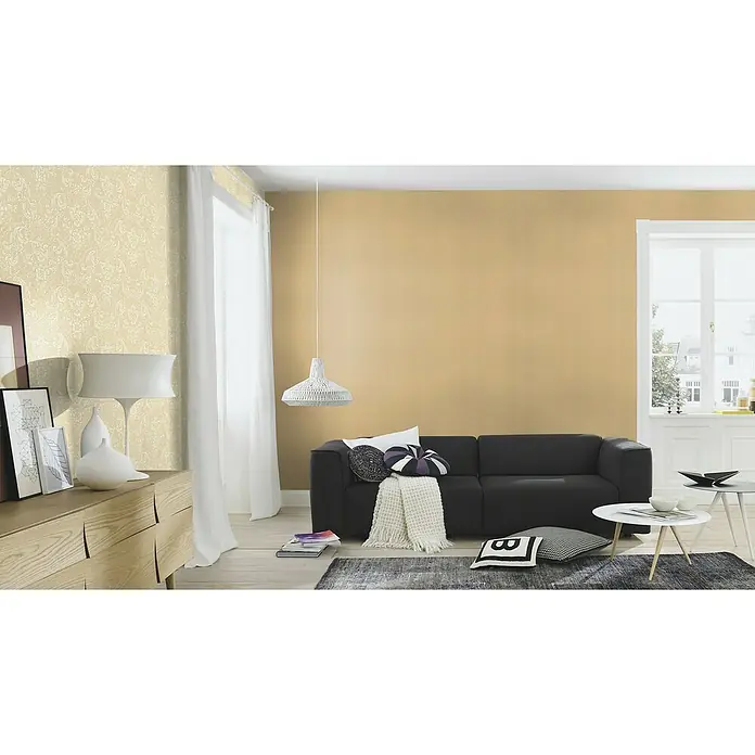 Rasch Trianon XIII Vliestapete Textil - Mattgold, Uni, 10,05 x 0,53 m - Wohnzimmer mit dunkelgrauem Sofa, Couchtisch und Holzkommode unter einer Lampe, die ein modernes und gemütliches Ambiente schafft.