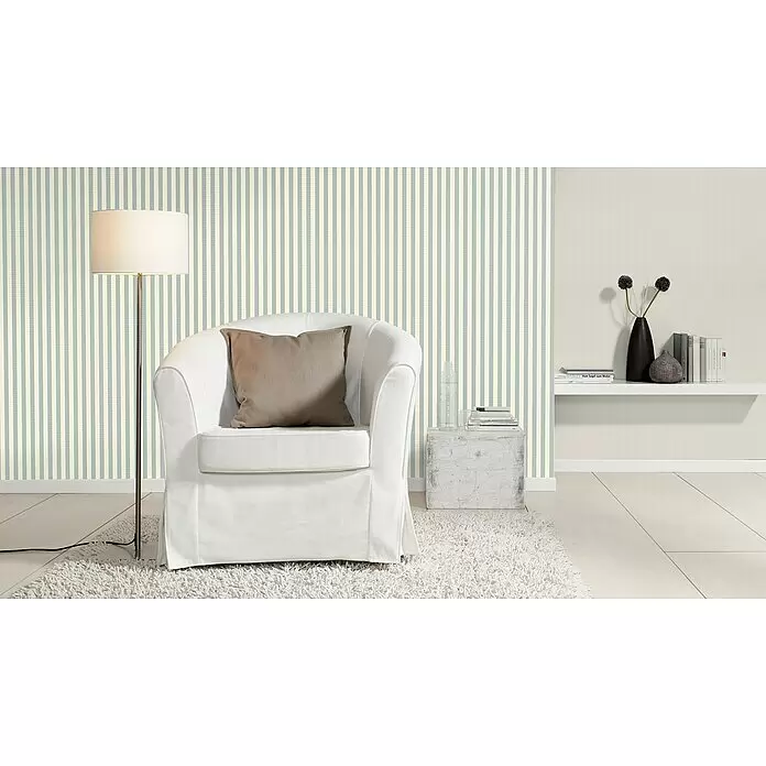 Rasch Trianon XIII Vliestapete - Blau-Weiß, Streifen, 10,05 x 0,53 m - Ein stilvolles Zimmer mit hellgrüner und cremefarbener gestreifter Tapete, ergänzt durch einen weißen Sessel, eine Stehlampe und ein minimalistisches Regal.