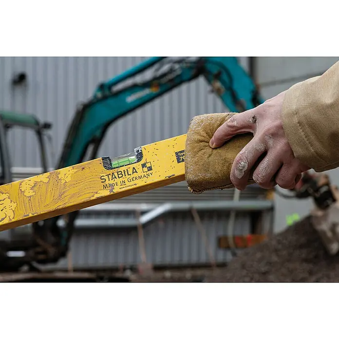 Stabila Wasserwaage Type 70Länge: 90 cm, Aluminium - Gelbe Stabila Wasserwaage wird mit einem Schwamm gereinigt, im Hintergrund ein Bagger. Präzisionswerkzeug "Made in Germany" für Handwerker.