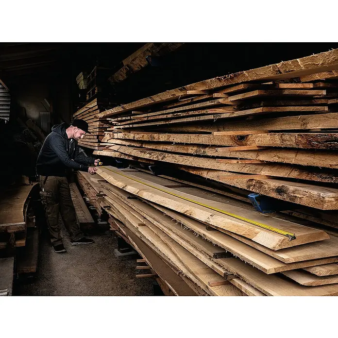 Stabila Taschenbandmaß BM 300 - 5 m - Ein Mann misst ein langes Holzbrett mit einem gelben Maßband in einer Holzwerkstatt, in der gestapelte Bretter lagern.