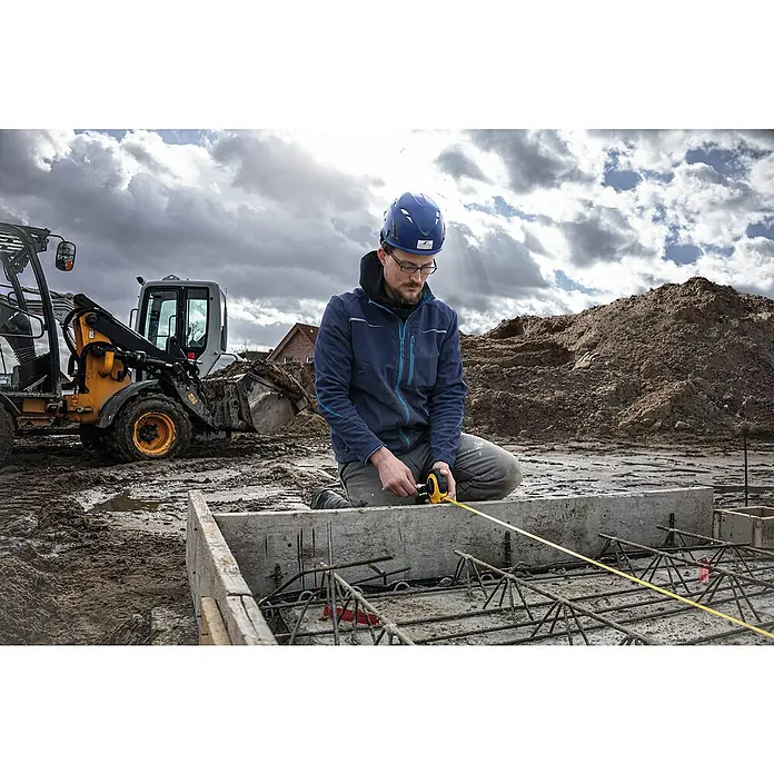 Stabila Kapselmaßband LBM 2000 Fibreglas20 m - Ein Bauarbeiter mit Schutzhelm misst mit einem gelben Messband eine Betonkonstruktion auf einer Baustelle, mit schwerem Gerät im Hintergrund.