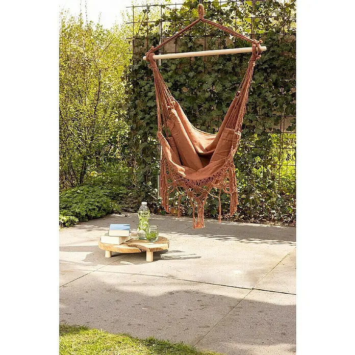 Hamaca individual Granada - L x An: 130 x 30 cm, Colores surtidos - Arreglo de jardín al aire libre con una hamaca marrón con flecos, una mesa de madera con decoración y plantas contra un exuberante fondo verde.
