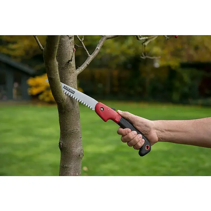 Kreator Serrucho de podar plegable Compact - Largo: 180 mm - Primer plano de una mano sujetando una sierra plegable Kreator roja y negra, recortando un árbol pequeño en un jardín.
