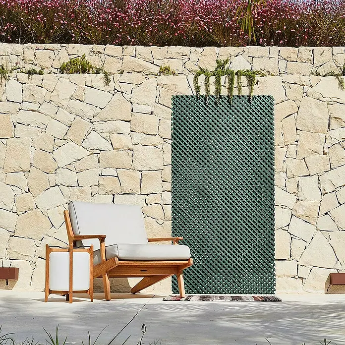 Celosía LOP - An x Al: 1 x 2 m, Verde - Escena exterior con una elegante silla de madera y mesa auxiliar frente a una pared de piedra decorativa.
