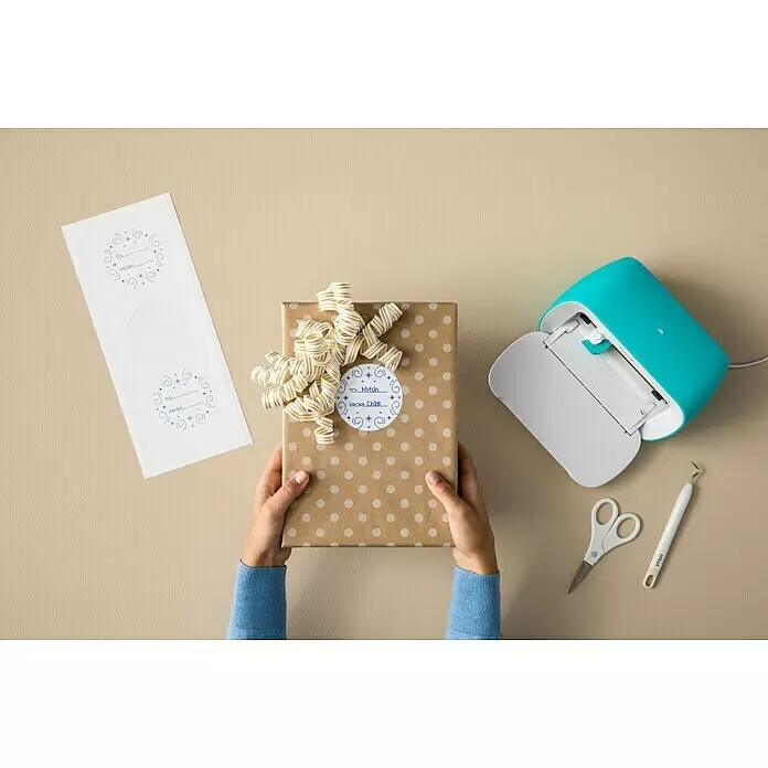 Cricut Joy Schneideplotter Smarte Schneide- und Zeichenmaschine - Türkis/Weiß, Bluetooth, Schnittbreite: 10,4 cm, Schnittlänge: 1,2 m - Eine türkisfarbene Cricut Joy Maschine mit Bastelwerkzeugen, Geschenkanhängern und einem gepunkteten Geschenk mit personalisiertem Anhänger.