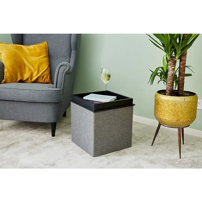 Store It Premium Sitzhocker - L x B x H: 37 x 37 x 41 cm, Grau - Ein grauer Pouf-Hocker mit schwarzem Tablett steht neben einem Sessel mit gelbem Kissen und einer Zimmerpflanze im goldenen Topf.