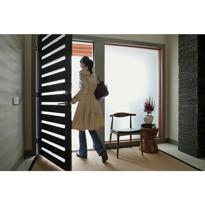 Ledvance Smart+ WiFi Bewegungsmelder - Weiß, L x B x H: 6,03 x 2,05 x 6,03 cm - Eine Frau betritt ein modernes Haus durch eine stilvolle Tür mit horizontalen Lamellen, während ein dekorativer Säule im Flur steht.