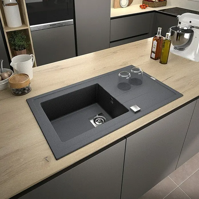 Carea Einbauspüle Tango - 86 x 50 cm, Betongrau, Material Spüle: CAREAlith® - Nahaufnahme einer modernen Küchenspüle mit Abtropffläche, die in eine helle Holzarbeitsplatte integriert ist. Sie ist umgeben von Küchenutensilien.