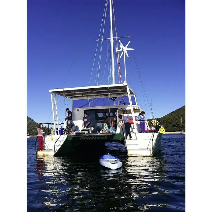 Sunset Windgenerator WG914 - 12 V, Max. Leistung: 300 W - Das Bild zeigt ein Segelboot mit einem Windrad und mehreren Personen an Bord, die eine Mahlzeit zubereiten, während ein Kajak im Wasser treibt.