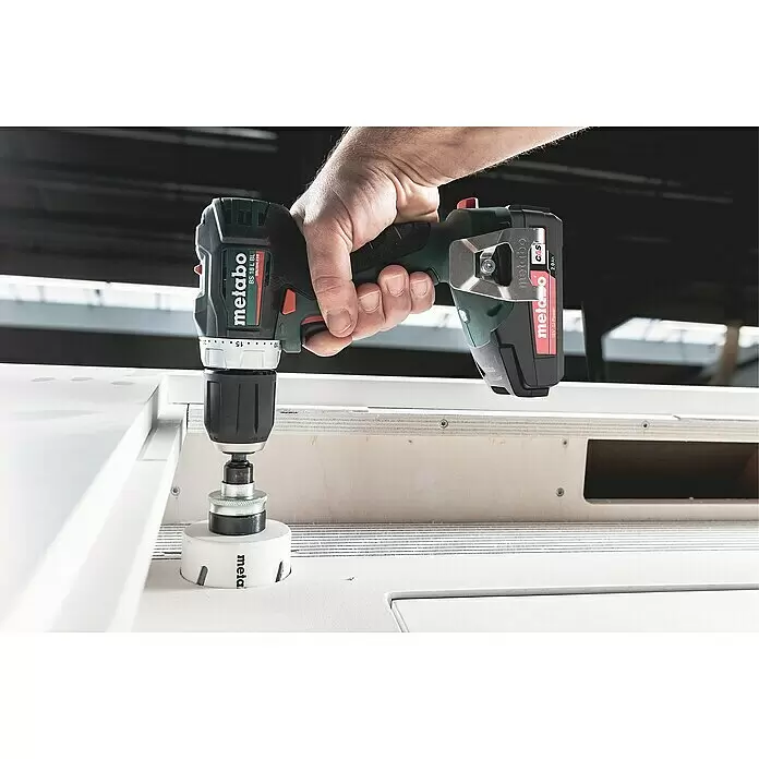 Metabo CAS 18V Akku-Bohrschrauber BS 18 L BL - 18 V, Ohne Akku, 60 Nm - Eine Hand bedient einen dunkelgrünen und schwarzen Metabo Akku-Bohrschrauber mit Lochsäge, die in eine weiße Platte schneidet.