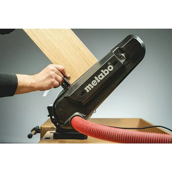 Metabo Band- & Tellerschleifer BS 200 Plus - 600 W, 2.980 U/min - Eine Metabo Kappsäge in Aktion, während ein Mann ein Holzbrett präzise zuschneidet. Der rote Staubabsaugungsschlauch ist ebenfalls zu sehen.
