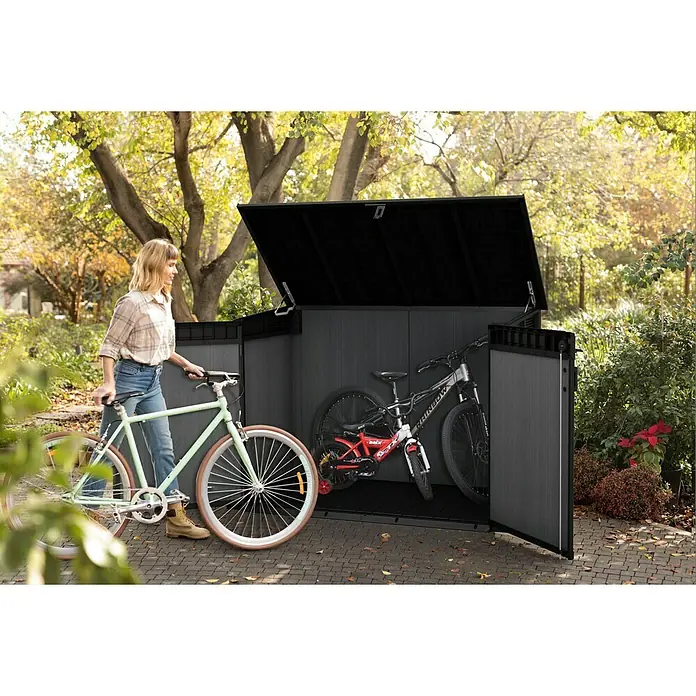 Keter Truhe Cortina Mega - Außenmaß inkl. Dachüberstand (B x T): 191 x 109 cm, Kunststoff, Anthrazit/Schwarz - Eine Frau schiebt ein mintfarbenes Fahrrad vor einen grauen Fahrradschuppen, in dem bereits Fahrräder stehen.