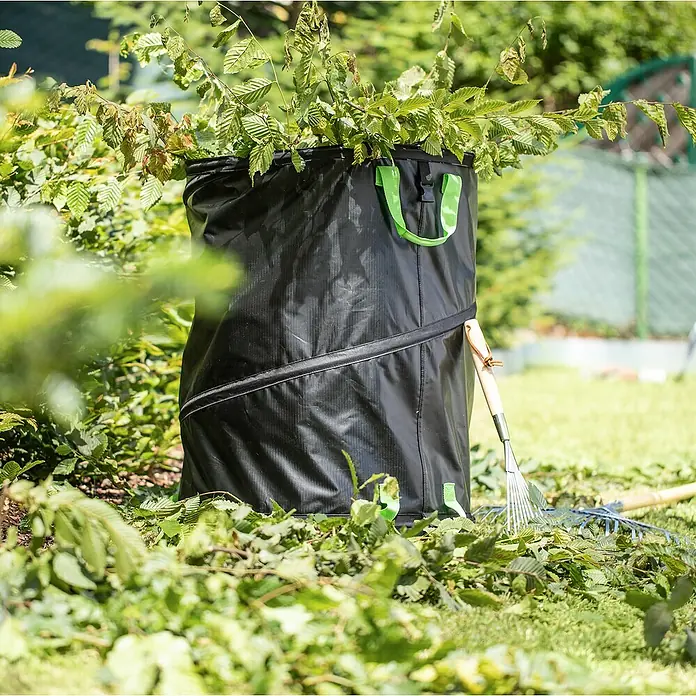 Windhager Pop-Up Gartenabfallsack Baglong-Life - Fassungsvermögen: 170 l, Ø x H: 56 x 70 cm, Schwarz - Gartensack gefüllt mit Blättern und Ästen, mit grünen Griffen und einem Rechen daneben. Perfekt für die Gartenarbeit!