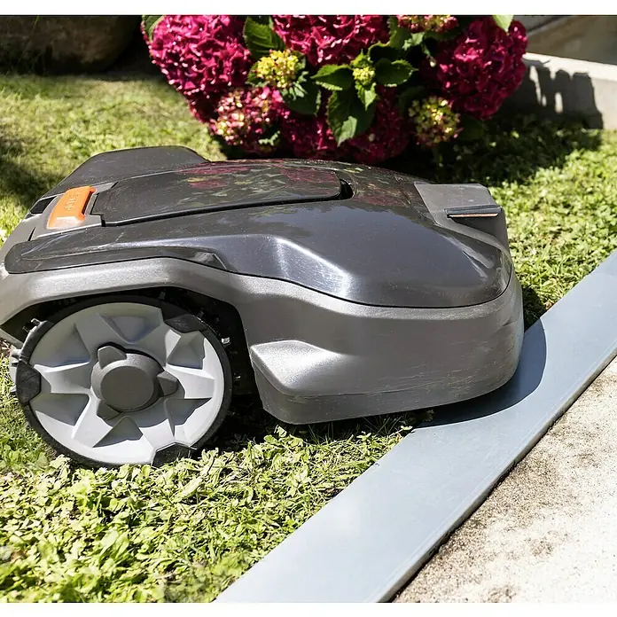 Windhager Mähkante Easymow - 80 x 11 x 7 cm, Silber, Verzinkt - Nahaufnahme eines grauen Rasenmähroboters auf Rasen mit bunten Blumen im Hintergrund. Der Rasenmäher steht neben einer grauen Kante.