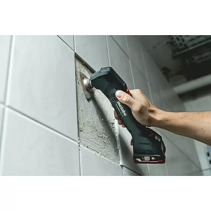 Metabo Akku-Multifunktionswerkzeug POWERMAXX MT 12 - 12 V, Ohne Akku - Eine Person verwendet ein dunkelgrün-schwarzes Metabo PowerMaxx MT 12 Multitool, um Material zwischen weißen Fliesen zu entfernen.
