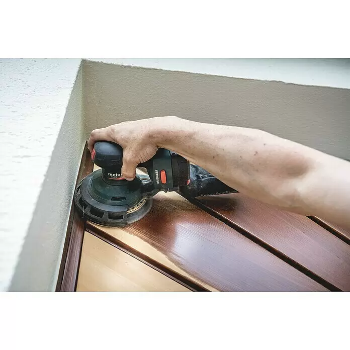 Metabo CAS 18V Akku-Exzenterschleifer SXA 18 LTX 150 BL - 18 V, Durchmesser Schleifteller: 150 mm - Hand hält einen grünen und schwarzen Exzenterschleifer, um eine Holzoberfläche neben einer hellen Wand zu glätten.