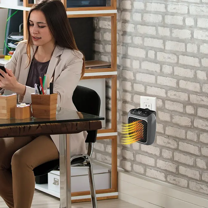 Calefactor cerámico Handy Heater - Gris, 11,5 x 14,6 x 22,9 cm - Una mujer está sentada en su escritorio mientras un calefactor compacto está enchufado en la pared, proporcionando calor.