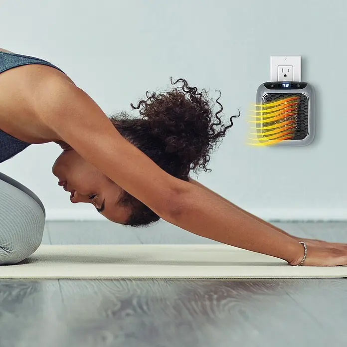Calefactor cerámico Handy Heater - Gris, 11,5 x 14,6 x 22,9 cm - Una mujer se estira sobre una esterilla de yoga junto a un calentador portátil enchufado a la pared.