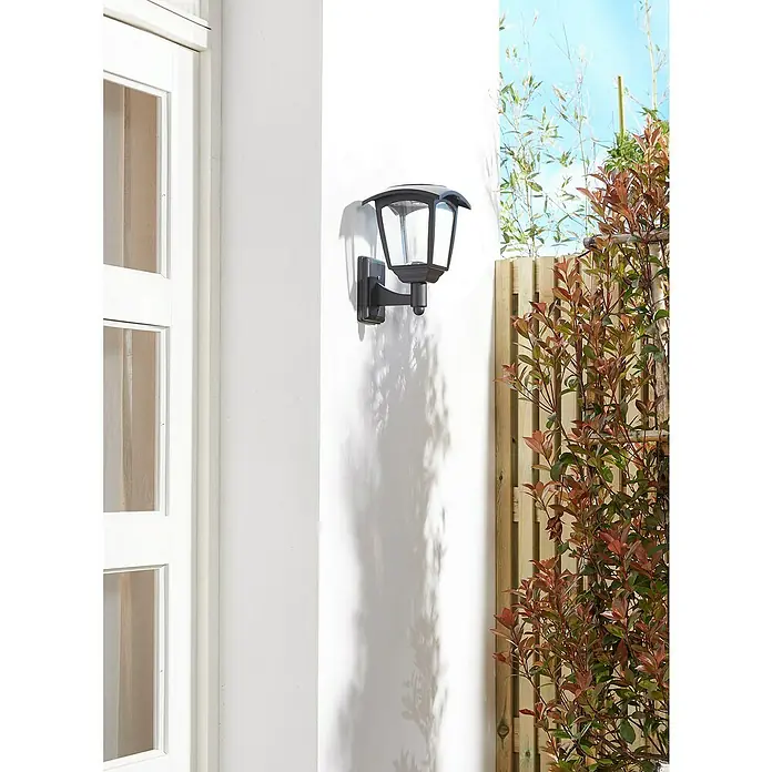 Luxform Minesota Aplique solar para exterior - L x An x Al: 20,8 x 18 x 26 cm, Negro, IP33 - Una elegante lámpara de pared negra con carcasa transparente, montada en una pared blanca junto a una ventana y plantas.
