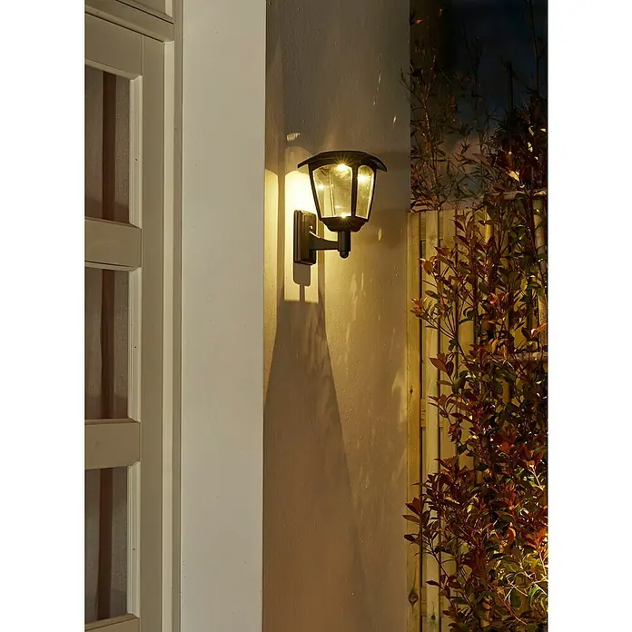 Luxform Minesota Aplique solar para exterior - L x An x Al: 20,8 x 18 x 26 cm, Negro, IP33 - Una lámpara de pared negra estilo farol ilumina una pared exterior, proporcionando iluminación ambiental en el jardín.