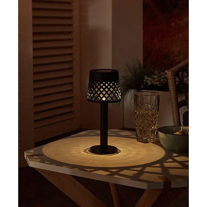 Luxform Manacor Lámpara de sobremesa solar - Negro, Ø x Al: 9 x 22 cm - Primer plano de una lámpara de mesa negra con un patrón geométrico sobre una mesa de exterior, que emite una luz cálida.