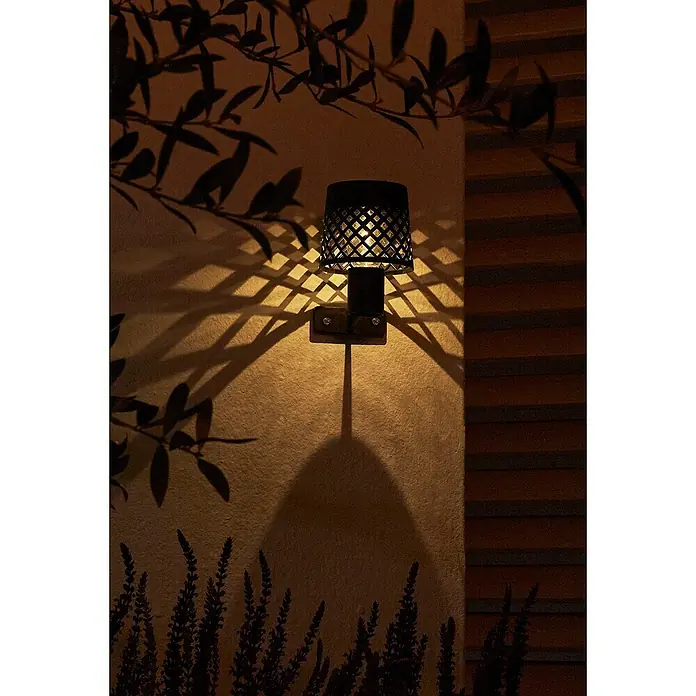 Luxform Manacor Aplique solar para exterior - L x An x Al: 9 x 9 x 13,5 cm, Negro - Primer plano de una lámpara de pared exterior negra con patrones geométricos que proyecta un brillo cálido en la pared, rodeada de sombras de plantas.