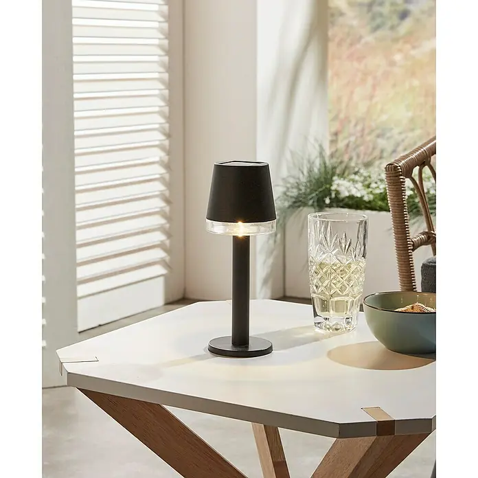 Luxform Cala D'Or Lámpara de sobremesa solar - Negro, IP44, Ø x Al: 8 x 22 cm - Primer plano de una elegante lámpara de mesa negra sobre una mesa blanca, junto a un vaso y un cuenco de aperitivos.