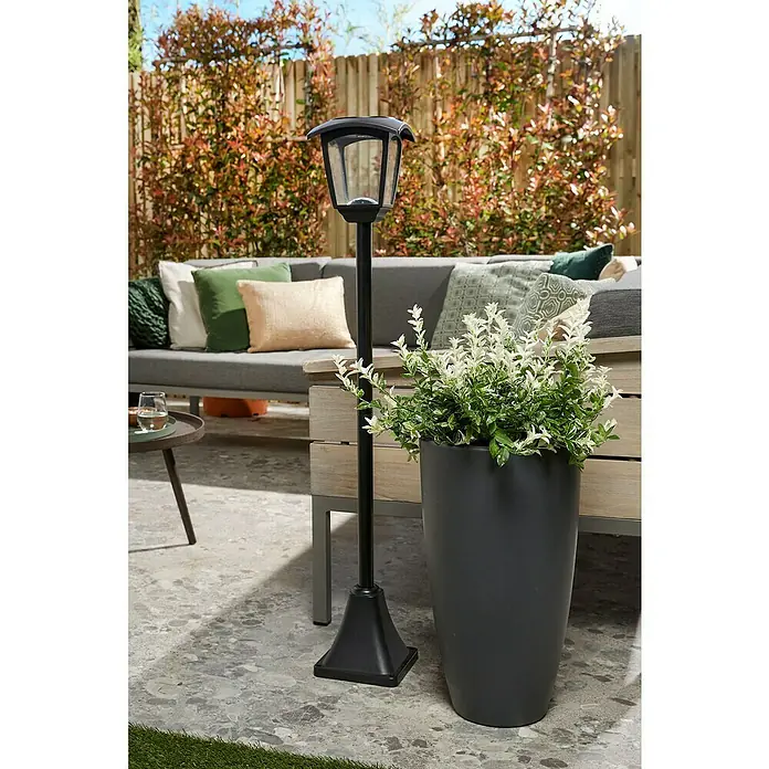 Luxform Minesota Farola LED Solar - Altura: 108,5 cm, Negro, IP33 - Una lámpara solar de jardín negra con cabeza de linterna y una gran jardinera con plantas blancas y verdes en una terraza.