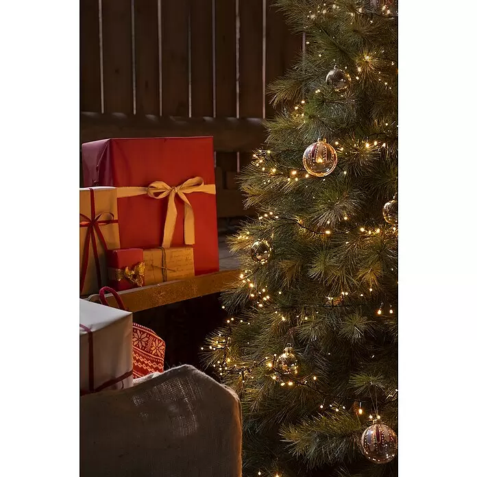 Konstsmide LED-Lichterkette Micro Compact - 11,78 m, Anzahl Leuchtmittel: 400 Stk., Netzbetrieben, 6 W - Ein festlicher Weihnachtsbaum mit warmen Lichtern und goldenen Kugeln neben verpackten Geschenken auf einem Holzregal und in einem Jutesack.