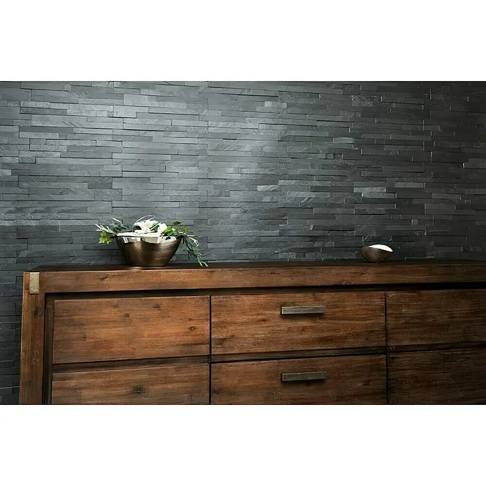 Verblendstein Nature - 15 x 60 cm, Schwarz, Steinoptik - Eine braune Holzkommode vor einer Wand mit grauen Steinpaneelen mit Deko-Elementen.