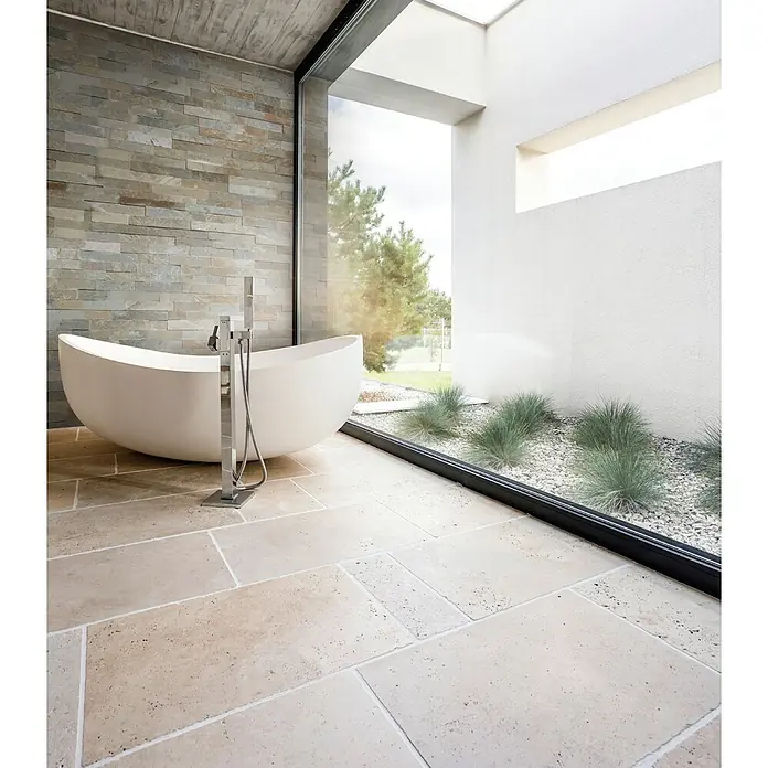 Verblendstein Nature - 15 x 60 cm, Beige, Steinoptik - Elegantes Badezimmer mit freistehender Badewanne, Natursteinwand und großem Fenster mit Blick auf einen Kiesgarten. Moderner Stil.