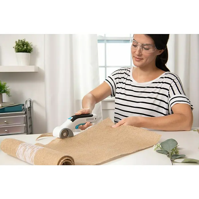 Black+Decker Cúter circular BCRC115-XJ - 3,6 V, 1,5 Ah, Iones de litio - Una mujer con gafas de seguridad utiliza un cortador rotatorio BLACK+DECKER para cortar arpillera sobre una mesa, con un rollo ya cortado.
