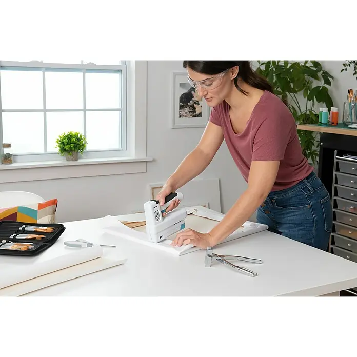 Black+Decker Grapadora de batería BCN115-XJ-Li - 3,6 V, 1 batería, 1,5 Ah - La mujer está utilizando una grapadora BLACK+DECKER para estirar el lienzo sobre un marco de madera, con herramientas y materiales a su alrededor.