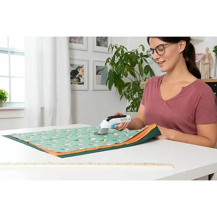 Black+Decker Grapadora de batería BCN115-XJ-Li - 3,6 V, 1 batería, 1,5 Ah - Una mujer usa un cúter rotatorio BLACK+DECKER para cortar tela sobre una mesa. La herramienta es blanca y azul con un mango negro.