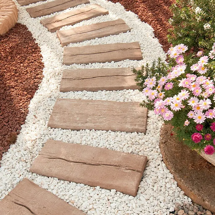 Traviesa de madera - 60 x 25 x 5 cm, Hormigón - Sendero de jardín con pasos de imitación madera incrustados en grava blanca, junto a piedras rojas y flores en floración. Los pasos forman un camino rústico.