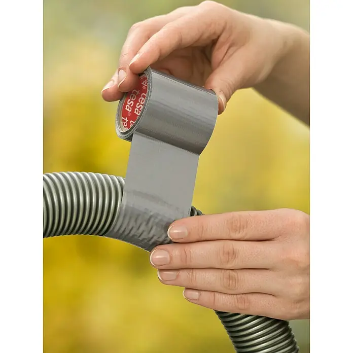 Tesa Cinta de reparación Extra Power - Gris, L x An: 5 m x 50 mm - Una mano está aplicando cinta adhesiva gris Tesa alrededor de una tubería acanalada gris para arreglarla. La cinta es fuerte y fiable.