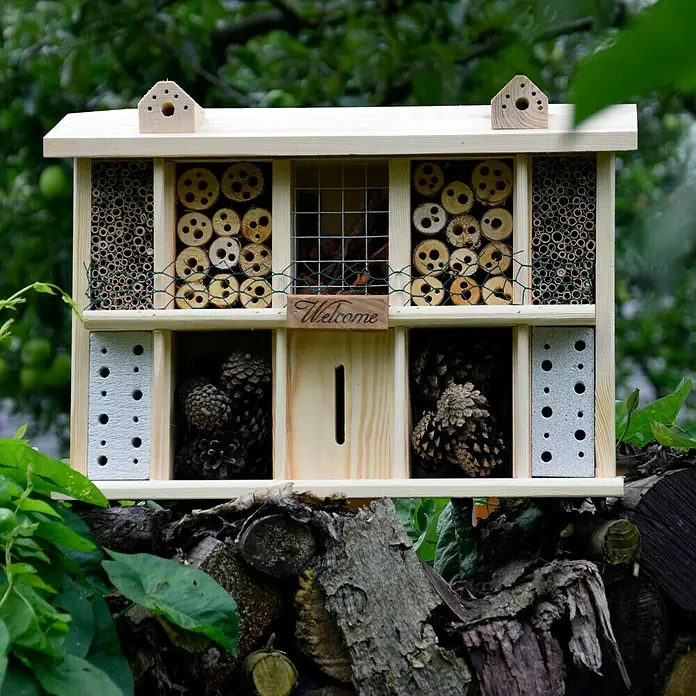 Dobar Insektenhotel Landsitz Superior - 47 x 12,5 x 34 cm, Kiefer - Holz-Insektenhotel mit verschiedenen Kammern für unterschiedliche Insektenarten, umgeben von Gartenbegrünung.