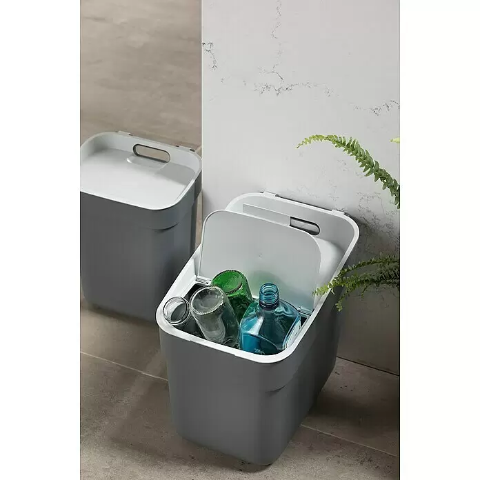 Curver Standard Mülleimer Ready to Collect - 20 l, Kunststoff, Dunkelgrau - Zwei graue Recyclingbehälter mit weißen Deckeln, einer geöffnet mit Glasflaschen darin, auf einem gefliesten Boden neben einer weißen Wand und einer Pflanze.