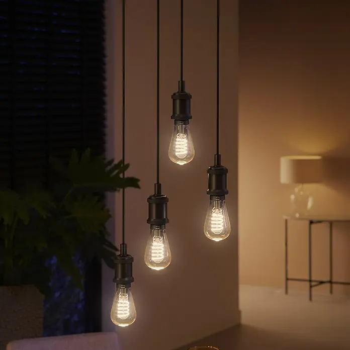 Philips Hue Smart-LED-Lampe White Ambiance Filament - E27, Dimmbar, Warmweiß, 550 lm, 7 W, Lampenbezeichnung: ST64, 1 Stk. - Eine Nahaufnahme von vier hängenden Edison-Glühbirnen, die ein warmes Licht in einem modernen Raum ausstrahlen.