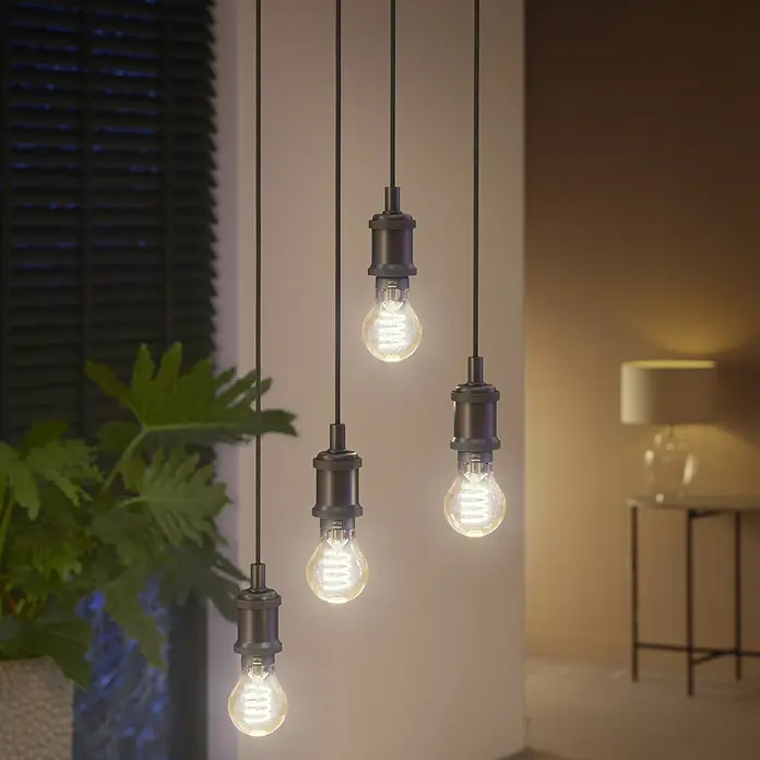 Philips Hue Smart-LED-Lampe White Ambiance Filament - E27, Dimmbar, Warmweiß, 550 lm, 7 W, Lampenbezeichnung: A60, 1 Stk. - Eine Nahaufnahme von vier Vintage-Glühbirnen mit Edison-Glühfäden, die von der Decke hängen und ein warmes Licht ausstrahlen.